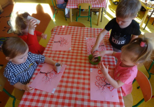 Czwórka dzieci stempluje rolkami papierowymi w kształcie serca koronę drzewa.